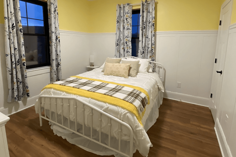 A cozy bedroom with a white bed, yellow accents, and floral curtains against a light yellow wall.
