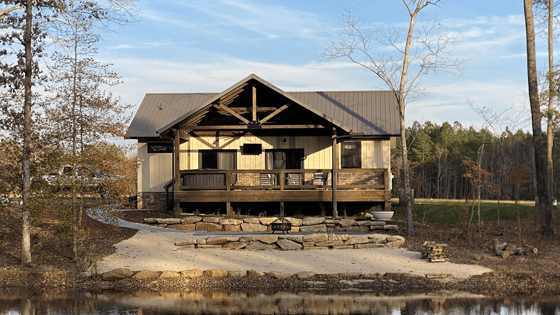 A cozy cabin with a porch overlooks a landscaped area by a pond.