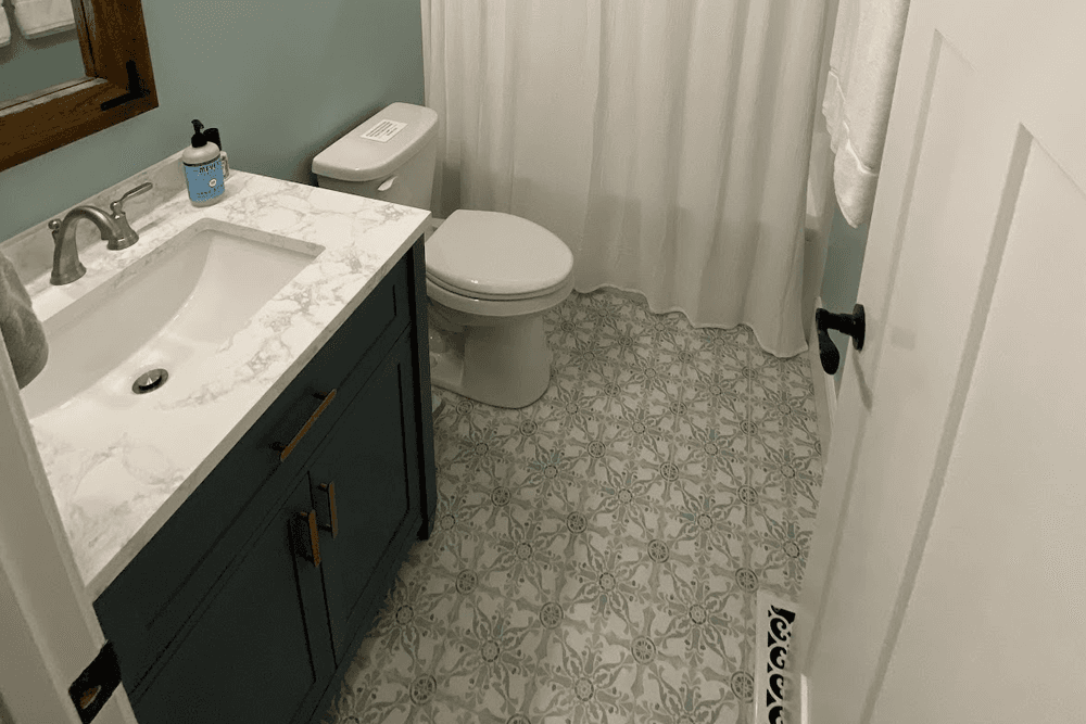 A small bathroom featuring a sink with a marble countertop, a toilet, patterned tile flooring, and light blue walls.